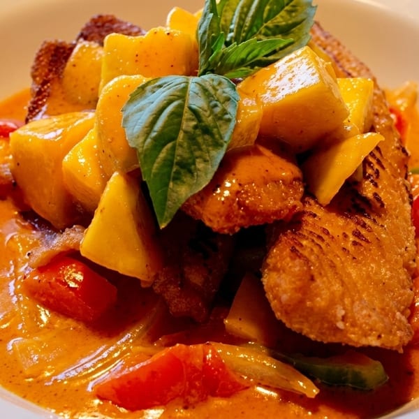 Crispy duck in red curry with mango, tomatoes, and basil in a white bowl.
